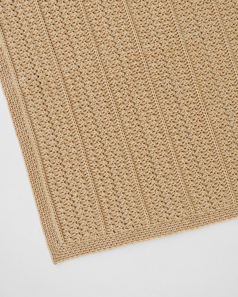 May include: A close-up of a tan crochet blanket. The blanket features a textured pattern with vertical lines and a detailed border. The neutral color and intricate design suggest a handmade item, suitable for home decor or as a cozy accessory.