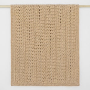 May include: A tan-colored, rectangular crochet blanket with a textured, vertical stripe pattern. The blanket is displayed hanging from a wooden rod against a white background. The edges have a solid border.