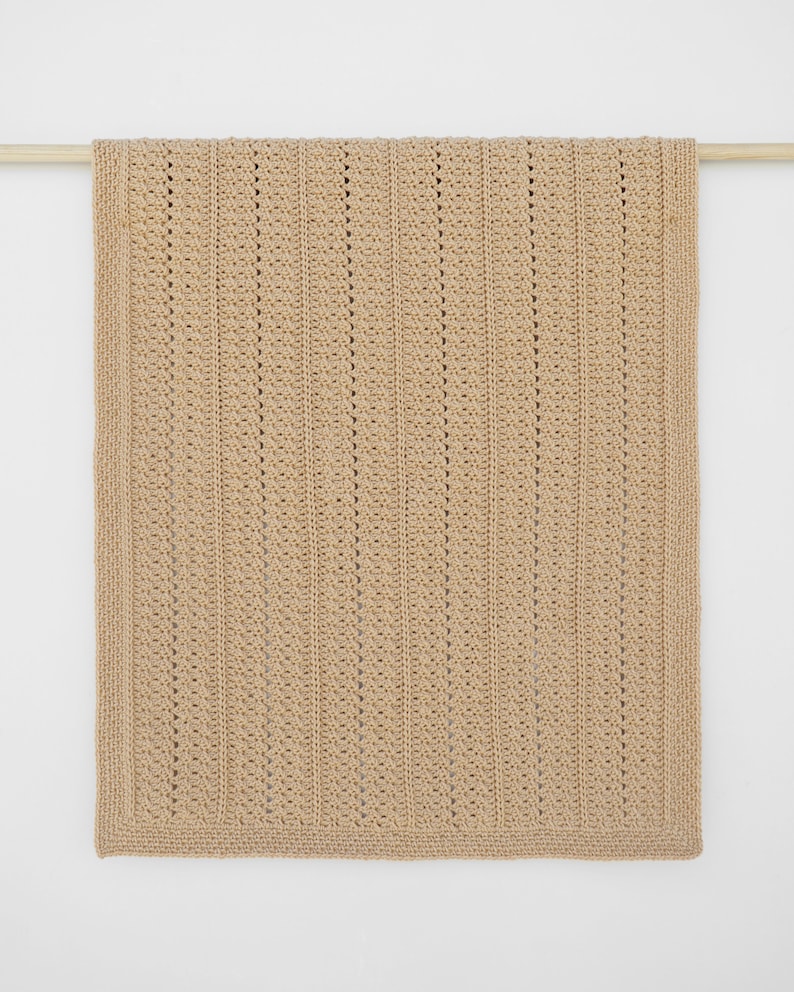 May include: A tan-colored, rectangular crochet blanket with a textured, vertical stripe pattern. The blanket is displayed hanging from a wooden rod against a white background. The edges have a solid border.