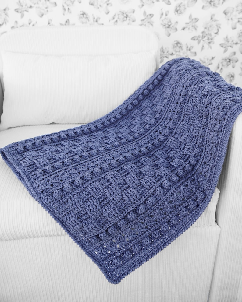 May include: A blue crocheted blanket with a textured pattern and a raised border. The blanket is draped over a white armchair.
