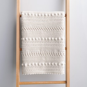 May include: A cream-coloured, hand-crocheted blanket with various textured patterns, including bobbles and diamond designs. The blanket is draped over a wooden ladder, showcasing its intricate details and craftsmanship. The blanket's dimensions are not visible.