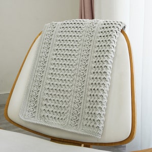 May include: A gray crocheted throw blanket draped over a white chair with wooden arms. The blanket has a textured pattern and a delicate border.