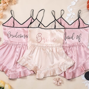 Puede incluir: Tres conjuntos de pijama de satén rosa con tirantes negros y ribete de encaje. Los conjuntos tienen diferentes textos en la parte delantera: "Bridesmaid", "Bride" y "Maid of Honor".