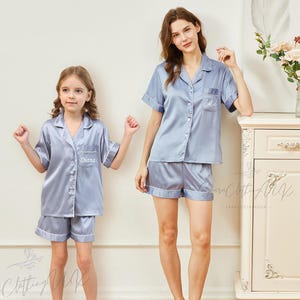 May include: Two people wearing matching light blue satin pajamas with short sleeves and shorts. The pajamas have white piping and are personalized with the names "Diana" and "Ellie".