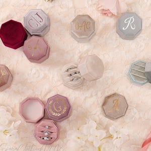 May include: A collection of velvet ring boxes in shades of burgundy, grey, and pink. Each octagonal box has gold lettering or initials. Some boxes are open, revealing rings inside.
