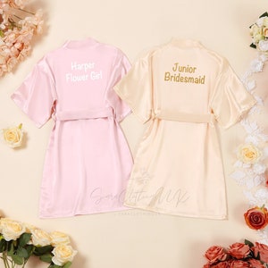 May include: Two satin robes, one pink and one cream, with embroidered text. The pink robe says "Harper Flower Girl" and the cream robe says "Junior Bridesmaid".