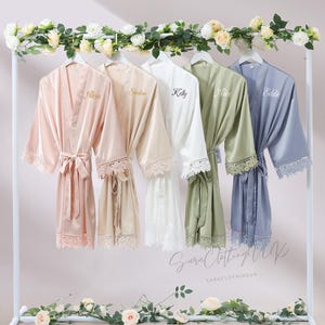 Könnte beinhalten: Fünf Satin-Bademäntel in Pastellfarben hängen an einem weißen Gestell. Jeder Bademantel, in den Farben Rosa, Beige, Weiß, Salbeigrün und Periwinkle, ist mit einem Namen und Spitzenbesatz versehen. Blumengirlanden schmücken das Gestell.