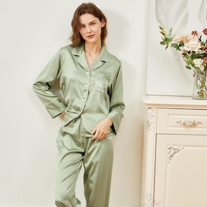 May include: A sage green satin pajama set with long sleeves and pants. The top has a button-up front and a pocket with the word "Claire" embroidered on it. The pants have a drawstring waist. The model is wearing white fuzzy slippers with the word "Bride" embroidered on them.