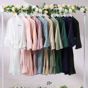 Peut inclure: Une présentation de peignoirs en satin colorés suspendus à un portant blanc, orné de guirlandes florales. Les peignoirs sont disponibles dans diverses teintes, dont blanc, rose, mauve, bleu, beige, vert, marine et noir. Chaque peignoir a de larges manches et une ceinture assortie.
