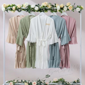Peut inclure: Cinq peignoirs en satin de différentes couleurs sont suspendus à un portant blanc orné de fleurs. Les peignoirs comprennent beige, vert, blanc avec "Bride" brodé, bleu-gris et rose. Chaque peignoir a une ceinture assortie et des manches à volants.