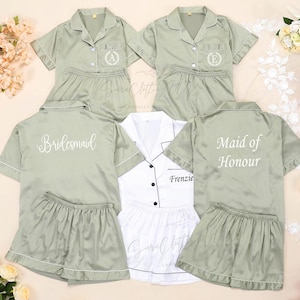 Könnte beinhalten: Salbeigrüne Satin-Pyjama-Sets mit weißem Paspel. Zwei Sets haben gestickte Initialen, eines sagt "Bridesmaid" und ein anderes "Maid of Honour". Ein weißes Pyjama-Set mit schwarzem Paspel ist ebenfalls enthalten. Blumengestecke umgeben die Sets.