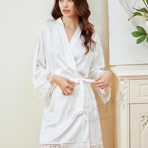May include: White satin robe with lace trim and a tie belt. The robe has a V-neck and long sleeves. The lace trim is on the sleeves and the bottom of the robe.