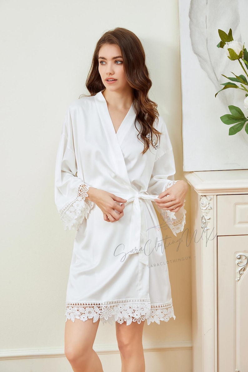 May include: White satin robe with lace trim and a tie belt. The robe has a V-neck and long sleeves. The lace trim is on the sleeves and the bottom of the robe.