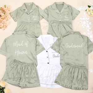 Puede incluir: Cuatro conjuntos de pijama de satén verde salvia con mangas cortas y pantalones cortos. Los conjuntos tienen texto blanco en la espalda que dice "Bride", "Maid of Honor", "Bridesmaid" y "Bride".