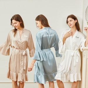 May include: Three satin robes in beige, blue, and white. Each robe has a ruffled hem, flared sleeves, and a matching belt. The beige robe has the name "love" embroidered on the front, the blue robe says "Bridesmaid" on the back, and the white robe says "Joyce".
