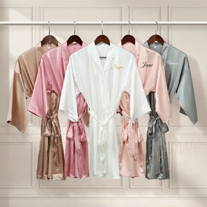 May include: Five satin robes hanging on a rack. The robes are in different colours: beige, pink, white, light pink, and grey. Each robe has a name embroidered on the front: Sara, Jane, Ariana.