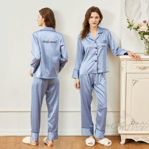 Könnte beinhalten: Hellblaues Satin-Pyjama-Set. Das langärmlige Oberteil hat schwarze Paspeln und Knöpfe vorne. Auf der Rückseite eines Oberteils ist das Wort "Bridesmaid" aufgedruckt, auf dem anderen der Name "Taylor". Die Hose hat eine passende schwarze Paspel.