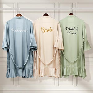 Puede incluir: Tres batas de sat&eacute;n con dobladillo con volantes, colgadas de perchas. Las batas son de color azul claro, beige y verde claro. La parte posterior de cada bata tiene una inscripci&oacute;n diferente: "Bride", "Bridesmaid" y "Maid of Honor".