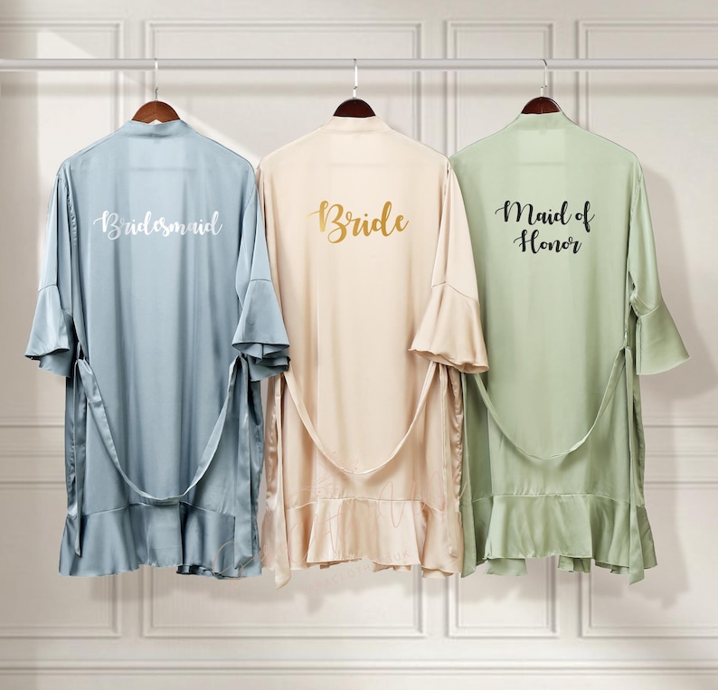 Puede incluir: Tres batas de sat&eacute;n con dobladillo con volantes, colgadas de perchas. Las batas son de color azul claro, beige y verde claro. La parte posterior de cada bata tiene una inscripci&oacute;n diferente: "Bride", "Bridesmaid" y "Maid of Honor".