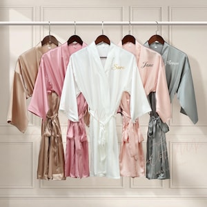 Puede incluir: Cinco batas de satén de varios colores cuelgan de una barra. Las batas son en tonos beige, rosa, blanco y gris. La bata blanca tiene el nombre "Sara" bordado en dorado. La bata rosa tiene el nombre "Jane" bordado en dorado, y la bata gris tiene el nombre "Ariana" bordado en dorado.