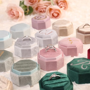 May include: An assortment of velvet ring boxes in various colors, including pink, green, and gray. Some boxes feature initials or a ring. The boxes are octagonal and have a soft, textured appearance, ideal for engagement rings.