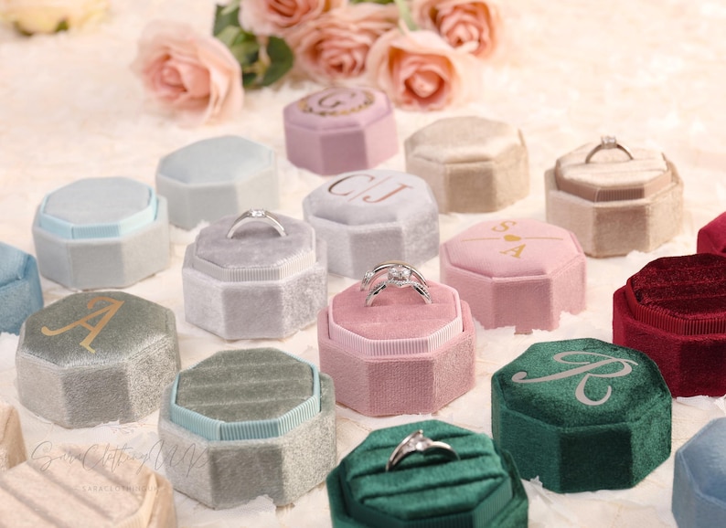 May include: An assortment of velvet ring boxes in various colors, including pink, green, and gray. Some boxes feature initials or a ring. The boxes are octagonal and have a soft, textured appearance, ideal for engagement rings.