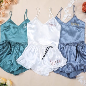 Puede incluir: Tres conjuntos de pijamas de satén a juego en verde azulado, blanco y azul claro. Cada conjunto incluye una camiseta sin mangas y pantalones cortos con un nombre personalizado bordado en la parte delantera. Los nombres son "Sam", "Grace" y "Emily".