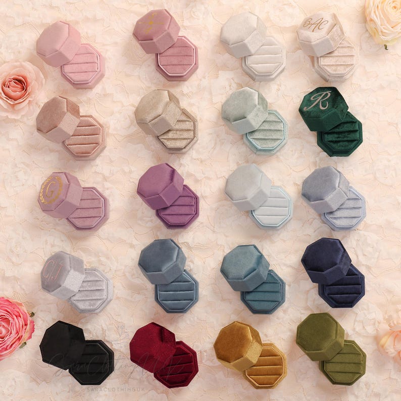 May include: An assortment of velvet ring boxes in various colors, including pink, green, and blue. Each box is octagonal and has a ribbed interior. Some boxes have initials embroidered on the lid.