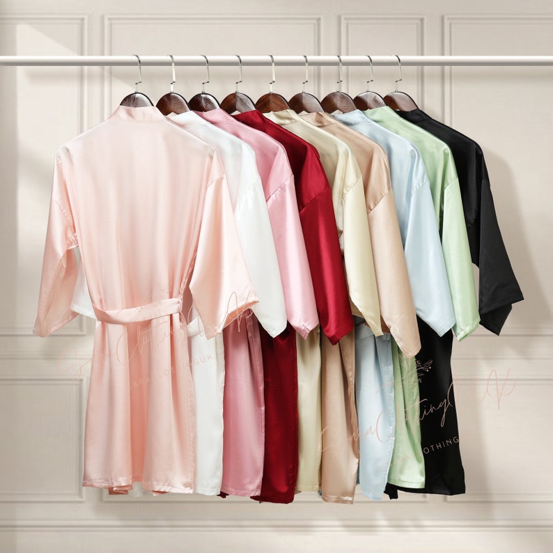 May include: A set of ten satin robes in various colours, including pink, white, red, beige, light blue, and green. The robes are hanging on hangers on a white wall.