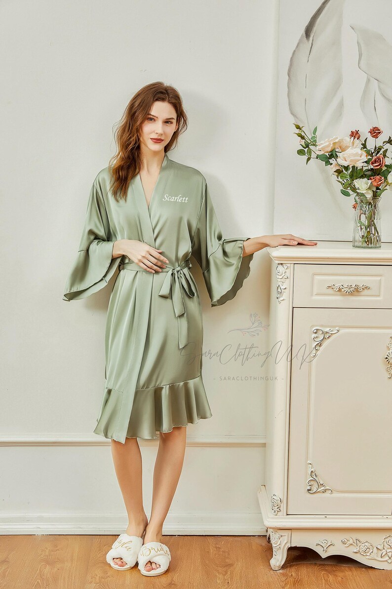 Puede incluir: Una bata de sat&eacute;n verde salvia con un dobladillo y mangas con volantes. La bata tiene un cintur&oacute;n de atar y est&aacute; bordada con el nombre "Scarlett". La modelo lleva zapatillas blancas con la palabra "Bride" bordada.