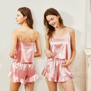 Puede incluir: Conjunto de pijama de satén rosa con top de tirantes y shorts. El top tiene tirantes finos y la palabra "Bridesmaid" o un nombre bordado. Los shorts tienen un dobladillo con volantes. Una opción de ropa de dormir cómoda.