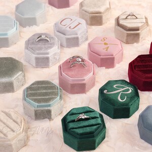 May include: An assortment of velvet ring boxes in various colors, including pink, green, and gray. Some boxes contain rings, while others feature initials or a monogram. The boxes are octagonal and have a soft, textured appearance.