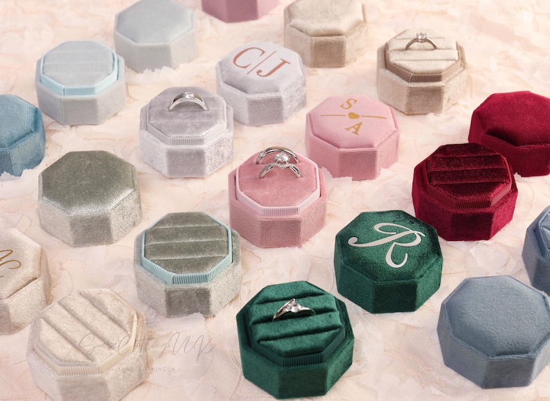 May include: An assortment of velvet ring boxes in various colors, including pink, green, and gray. Some boxes contain rings, while others feature initials or a monogram. The boxes are octagonal and have a soft, textured appearance.