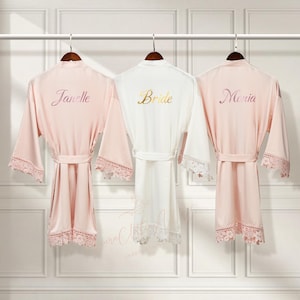 May include: Three satin robes with lace trim, hanging on hangers. The robes are pink, white, and pink. The robes are embroidered with the names "Janelle", "Bride", and "Moria".