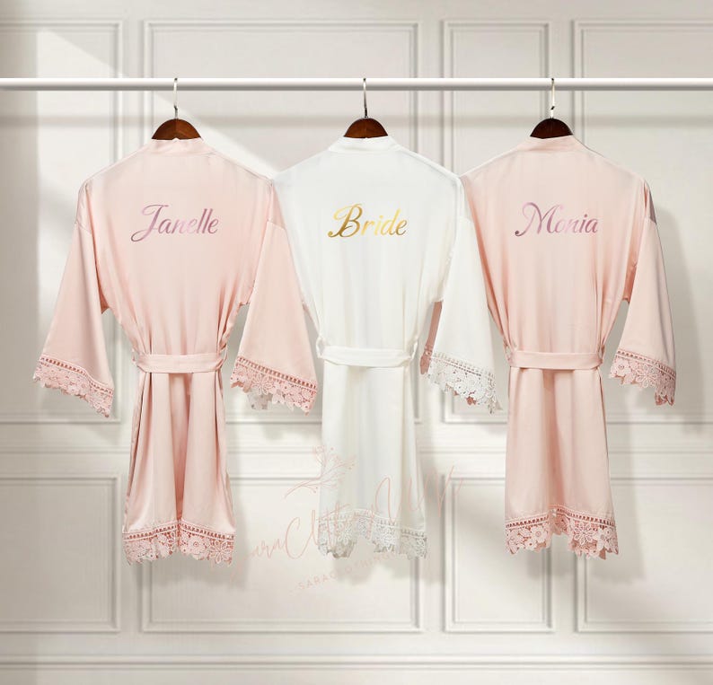 May include: Three satin robes with lace trim, hanging on hangers. The robes are pink, white, and pink. The robes are embroidered with the names "Janelle", "Bride", and "Moria".