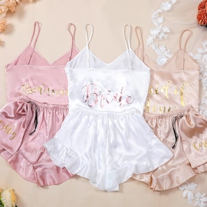 Puede incluir: Tres conjuntos de pijama de satén con letras doradas. El conjunto rosa dice "Bridesmaid" y "Ashley", el conjunto blanco dice "Bride" y el conjunto melocotón dice "Maid of Honor" y "Corinna".