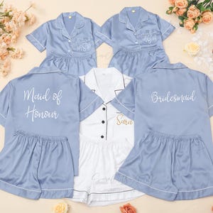 Puede incluir: Conjuntos de pijama de satén azul claro con ribete blanco y texto personalizado. Un conjunto es blanco con ribete negro. El texto incluye "Maid of Honour", "Bridesmaid", iniciales y nombres. Arreglo floral en el fondo.
