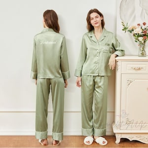 Könnte beinhalten: Zwei Frauen in passenden salbeigrünen Satin-Pyjama-Sets. Die Pyjamas bestehen aus einem Hemd mit Knöpfen und Brusttasche sowie einer Hose. Auf einem Set steht "Bridesmaid" auf dem Rücken. Auf dem anderen ist ein Name auf der Tasche gestickt.