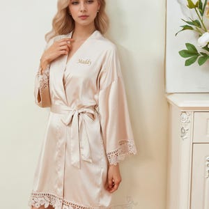 May include: A light beige satin robe with a lace trim. The robe has a tie belt and is embroidered with the name "Maddy".