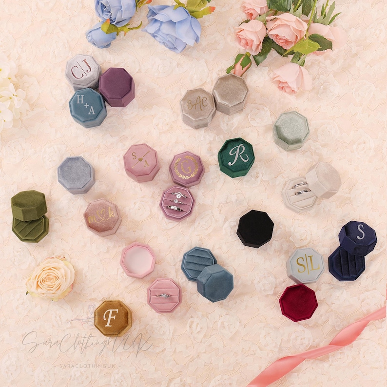 May include: An assortment of velvet ring boxes in various colors, including green, pink, and blue, are arranged on a textured surface. Some boxes are open, displaying rings. Several boxes have initials or monograms on the lids. Flowers and a ribbon are also present.