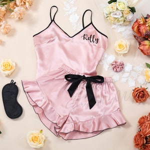 Puede incluir: Un conjunto de pijama de satén rosa con ribetes negros y un lazo de satén negro. La parte superior tiene cuello en V y los pantalones cortos tienen volantes. La parte superior tiene el nombre "Kelly" bordado en la parte delantera. El conjunto se acompaña de una máscara para dormir negra.