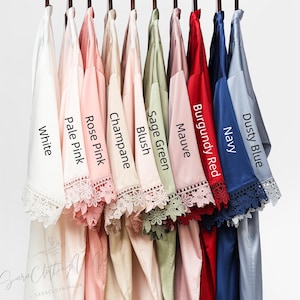 May include: Ten satin robes in different colors, hanging on a rack. The robes have lace trim and are labeled with the following colors: White, Pale Pink, Rose Pink, Champagne, Blush, Sage Green, Mauve, Burgundy Red, Navy, Dusty Blue.