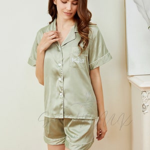 May include: A sage green satin pajama set with short sleeves and shorts. The top has a white button closure and a pocket with the name "Kita" embroidered on it.