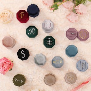 May include: An assortment of velvet ring boxes in various colors, including burgundy, navy, green, and gray. Some boxes feature gold initials or a monogram, while others hold engagement rings. The boxes are octagonal and arranged on a cream-colored surface.