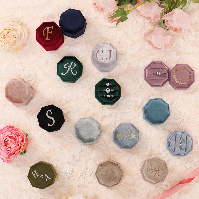 May include: An assortment of velvet ring boxes in various colors, including burgundy, navy, green, and gray. Some boxes feature gold initials or a monogram, while others hold engagement rings. The boxes are octagonal and arranged on a cream-colored surface.