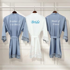 May include: Three satin robes with lace trim, hanging on hangers. The robes are in blue, white, and blue. The robes are embroidered with the text "Mother of the Bride", "Bride", and "Maid of Honor".