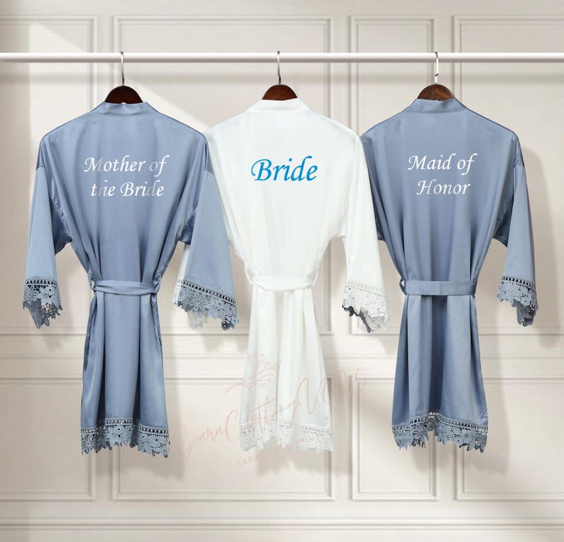 May include: Three satin robes with lace trim, hanging on hangers. The robes are in blue, white, and blue. The robes are embroidered with the text "Mother of the Bride", "Bride", and "Maid of Honor".