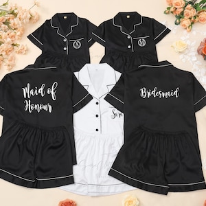 Pode incluir: Conjuntos de pijama de cetim preto com debrum branco e bordados personalizados. Um conjunto diz "Maid of Honour", outro "Bridesmaid", e outros têm iniciais. Um conjunto branco também está incluído.