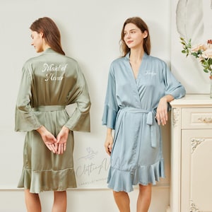 May include: Two women wearing satin robes. One robe is sage green with "Maid of Honor" printed on the back. The other robe is light blue with a name printed on the front. Both robes have a belt and ruffled hem.