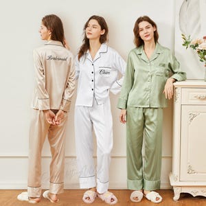 May include: Three sets of satin pajamas in beige, white, and sage green. Each set includes a button-down shirt and pants with black trim. The beige shirt has "Bridesmaid" embroidered on the back, while the white shirt has "Claire" embroidered on the front.